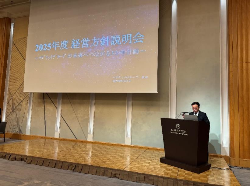『2025年度 経営方針説明会』のタイトルスライドが映し出されたスクリーン前で、演台に立つスーツ姿の男性が発表している様子。