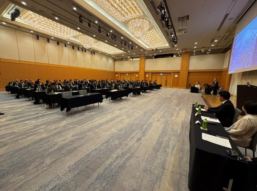 ホテルの大宴会場で行われた経営方針説明会。スーツ姿の参加者が整然と並び、壇上では発表者がスライドを映して説明している様子。